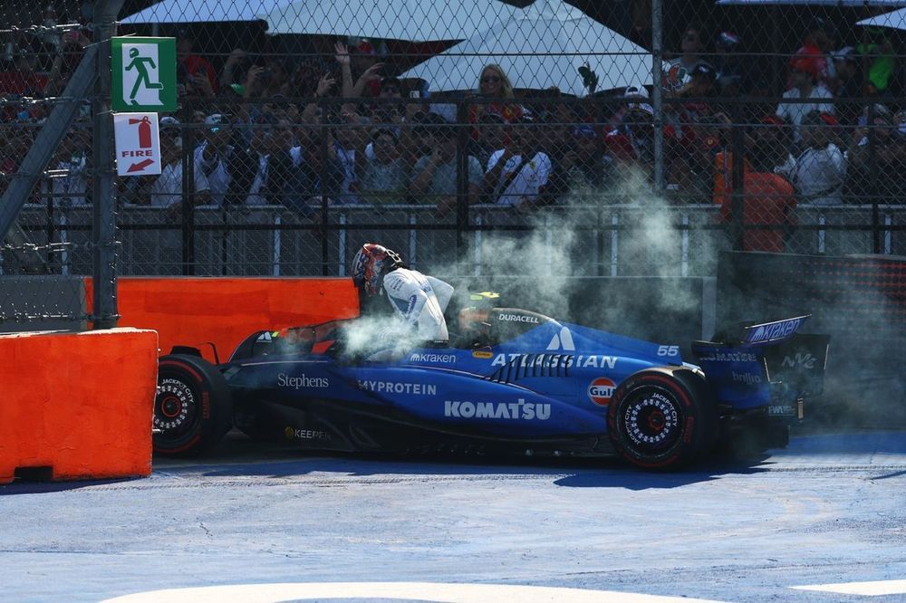 A picture of a Williams Formula 1 car from the side. Smoke is coming out and the driver is getting out of the car. The car crucially is not behind a barrier