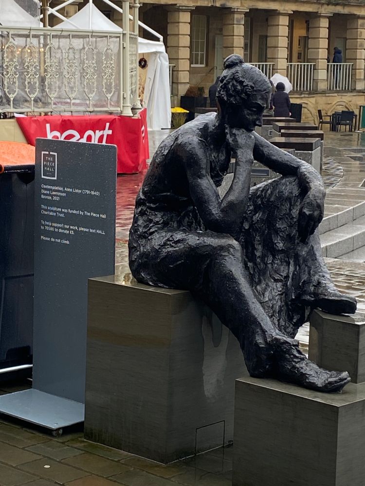 A very sad and wet looking photo of the Anne Lister statue at the Piece Hall in Halifax. The statue is of Anne Lister, who is sat down with her chin resting on her fist, looking pensive. 