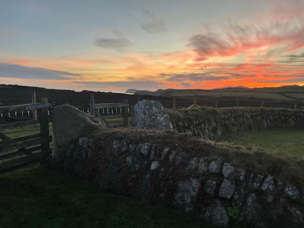 A stone wall with wooden gate in the foreground with a vivid sunset above a sliver is sea. 