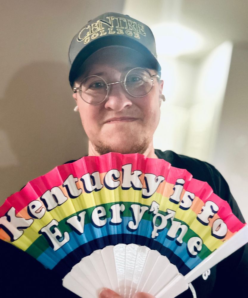A pic of the OP, a white trans masculine person with glasses, smiles at the camera. They are wearing a Centre College baseball cap and holding up a fan in Pride colors which has the text, “Kentucky is for Everyone”.