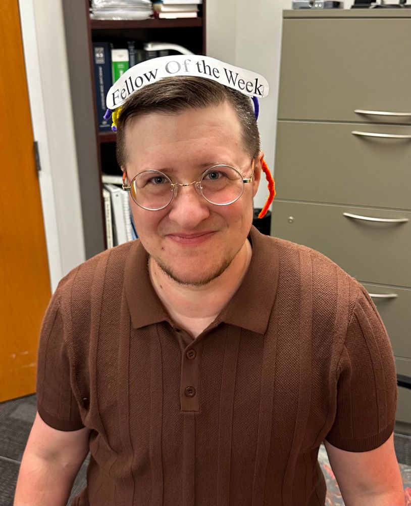 A picture of a white masculine looking person with brown hair and silver circular glasses. They are wearing a brown polo and a pipe cleaner crown with the words, “Fellow of the Week”.