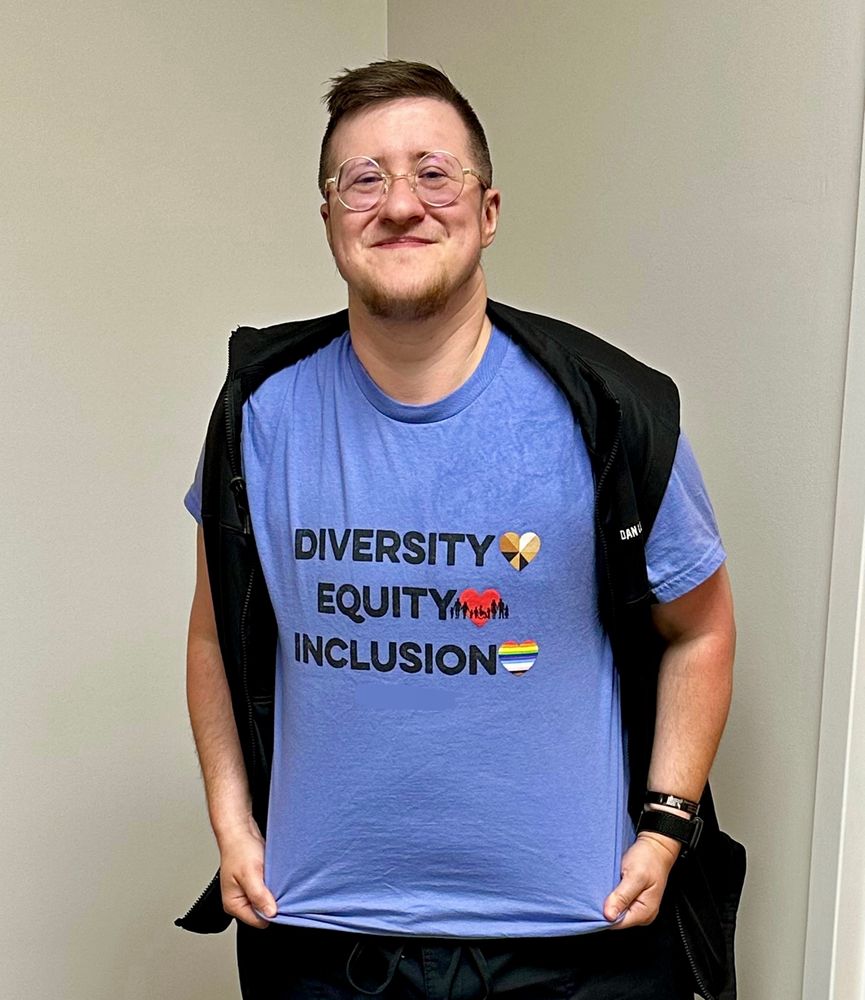 A picture of a white masculine looking person with circular glasses and short brown hair. They are wearing a light blue shirt which has the words, “Diversity, Equity, Inclusion”.