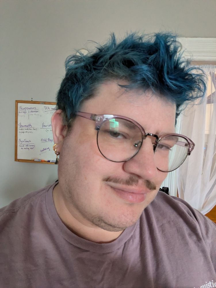 kirby selfie: a white nonbinary with blue hair, glasses, mustache, and gay little earrings