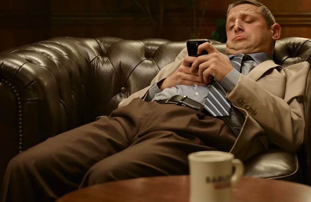 Tim Robinson slumped very far down in a big chair looking at his phone.