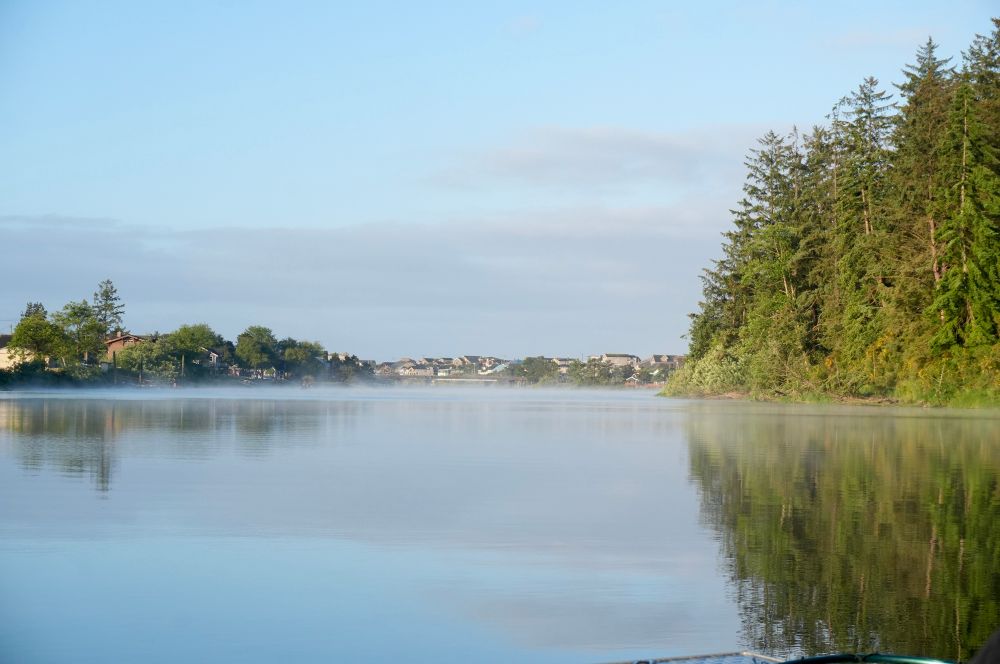 Landscape photo of the Nestucca River looking toward Pacific City. The river is calm and glassy showing reflections of trees that line the riverbank
