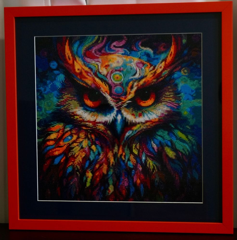 Photo of framed cross-stitch portrait of a multi-colored owl. Owl feathers contain different blues, greens, oranges, and reds. 