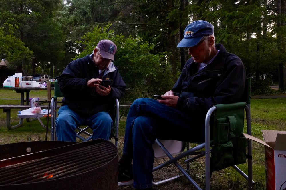 Photo of two 80ish year old men sitting in camp chairs around a campfire at Devil’s Lake Campground staring at their phones