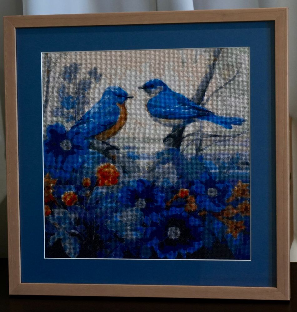 Photo of a framed cross-stitch portrait of blue birds perched on branches with red, blue and yellow/orange flowers in foreground and trees in background. 