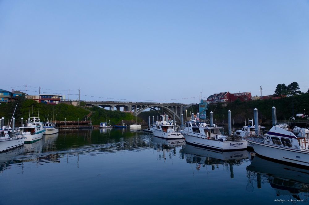 Photo of the harbor at Depoe Bay, Oregon. The bay is calm and the water is smooth. Charter fishing boats line the docks, waiting to fill up with anglers and head out into much rougher ocean conditions (on this day the swells were 6-8 feet). The Depoe Bay Bridge can be seen in the background. 
