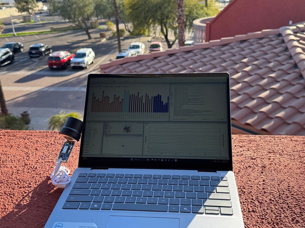 A laptop resting outdoors on a ledge with a bare circuit board global navigation satellite system receiver connected to an antenna plugged in via the left side of the computer. The laptop’s screen is displaying various types of information related to global navigation satellite systems, such as which satellites are in view of the receiver. 