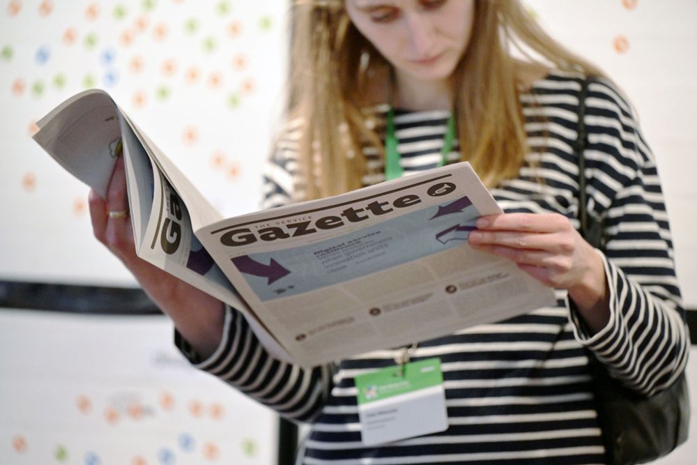 A woman with long hair, a stripy longsleeve shirt and a lanyard holding a newspaper reading it; its masthead says ‘The Service Gazette’; the headline next to several arrows says “Digital service resilience”