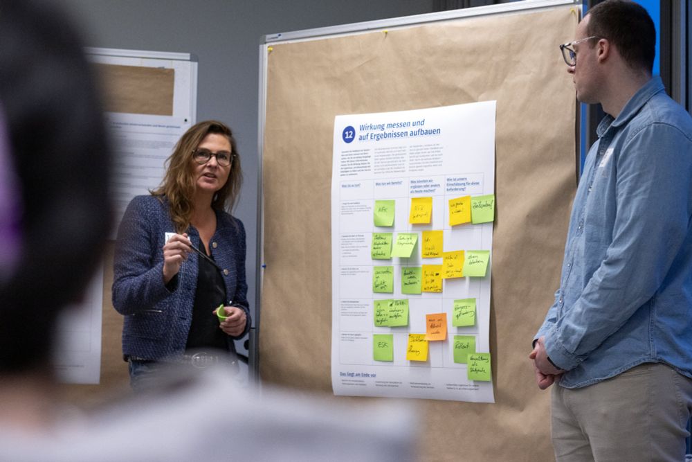 2 people, a woman and a man, standing in front of a mobile wall with a poster with various sticky notes in green, yellow and orange, headlined “12, measuring impact and working with results”, the woman is speaking, holding a pen and note pad, a person in the foreground seems to be listening