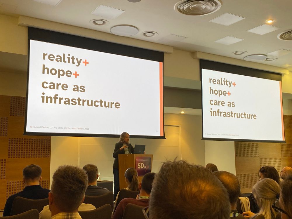 A woman with light skin and grey hair standing at a lectern in front of an audience with a projected slide above her saying: “reality + hope + care as infrastructure”