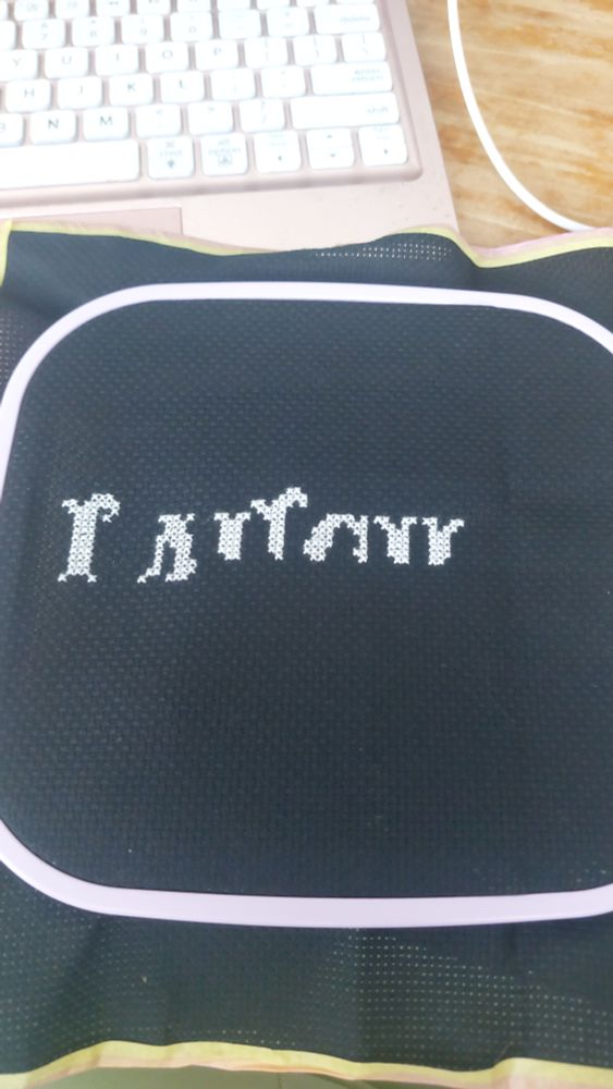 Een zwart borduur lapje in een lila borduurring gespannen. Er is geborduurd met lichtgrijs garen, maar het is niet goed te zien wat het moet voorstellen