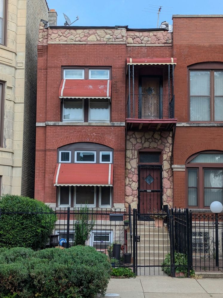 Color photo of a raised two-story red-brick row-house, with bay for windows on left and door/balcony on right. Brick plain, with gently rounded arch over first-floor windows and flat arch over second-floor windows. Metal awnings, red with white stripes at ends, below transoms on both windows and on second-floor balcony. Permastone in tan and pink around front door and on parapet. 