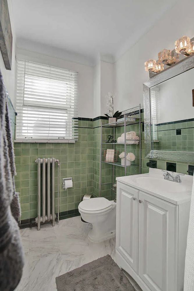 Color pic of bathroom. Bottom 2/3rd of walls are clad in medium-sized square green tiles, with slight variations in color. Black tiles at the edges, including pencil tiles below the top row of green tiles. Newer marble-y floor and sink.