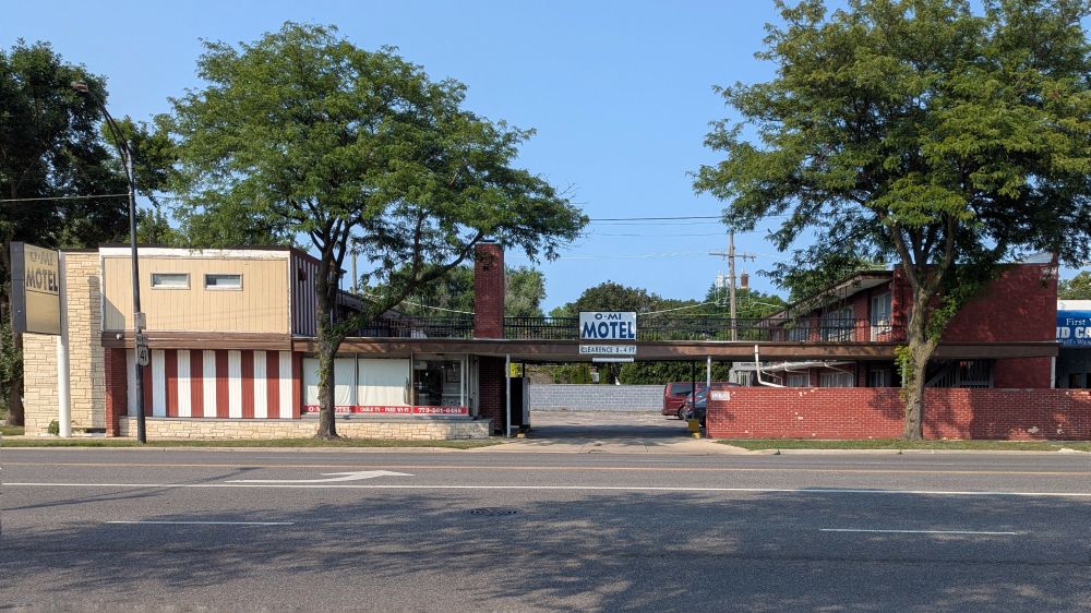 Color photo of a long two-story motel, in red brick, faux stone, and wood or similar. Rooms in two tiers across a parking lot. Walkway bridge over lot entrance, with sign: O-MI MOTEL Clearence 8-4 ft.