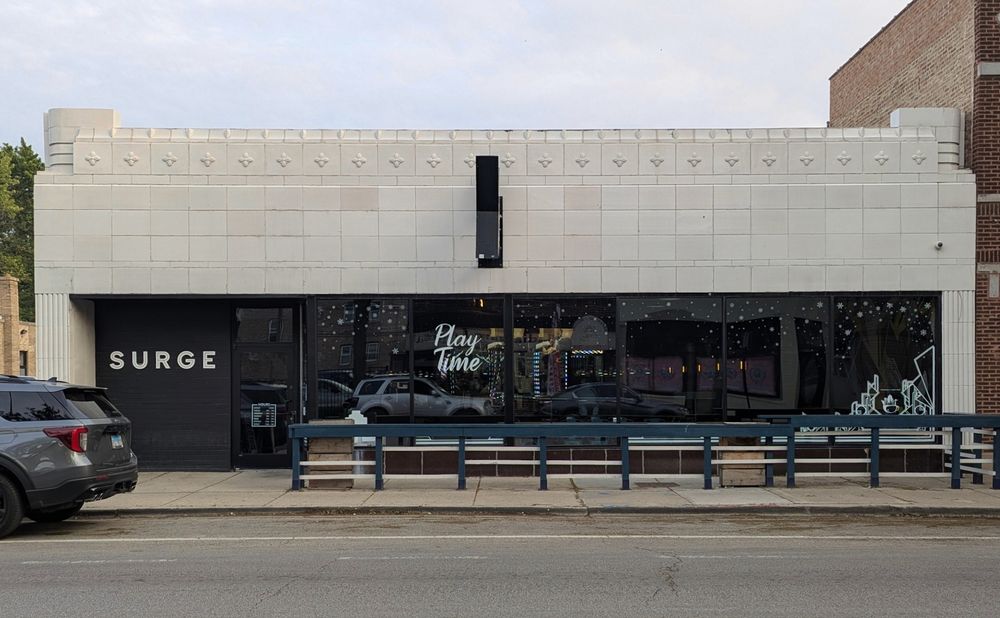 Color photo of a 1-story retail building clad in white terra cotta under a smokey sky. Large windows at street level. Most of the facade is plain white blocks, with fluted pilasters at the edges. The parapet has slightly more decorated blocks with diamond shapes in the center and blocks at the end with a curve at the corner. A sign by the door says SURGE. On the windows: "Play Time." 