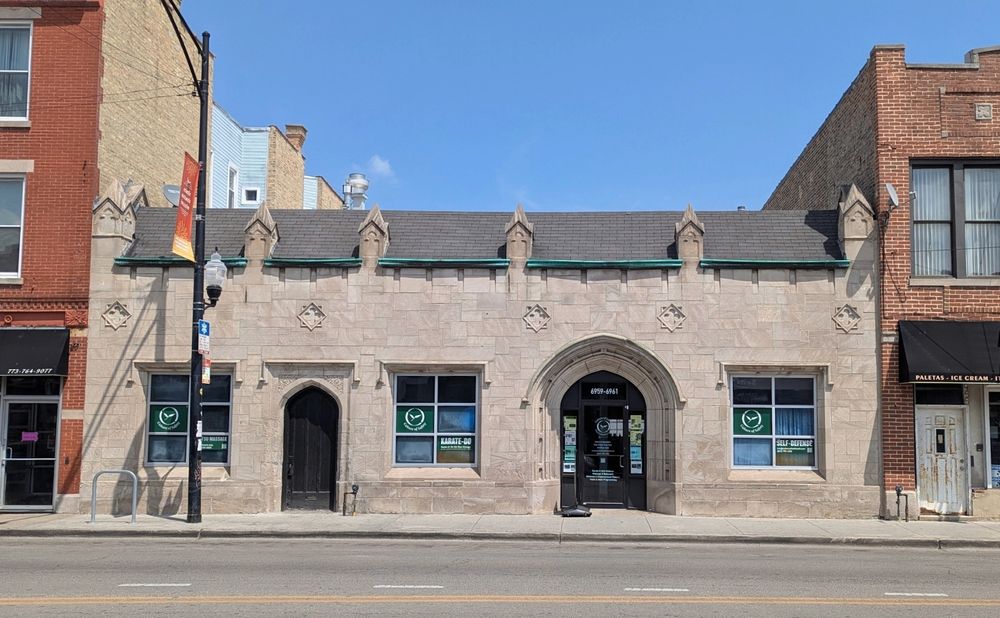 Color photo of a one-story commercial building, wedged between taller building. Limestone or facsimile light grey blocks in broken course. Stone punches through roofline six times, ending in pointy chunky finials. Main entrance is gently pointed compound arch, smaller door on the left. Three sets of store windows. Between the doors/windows and the parapet a design of quatrefoils within diamonds over squares.