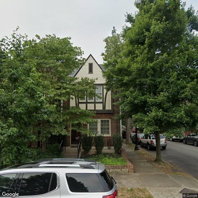 Street View of a two-story Tudor Revival home in brown brick, with cross-gable featuring combination of brown on tan half-timbering and white siding. Steps up to porch. Trees partially obscure view.