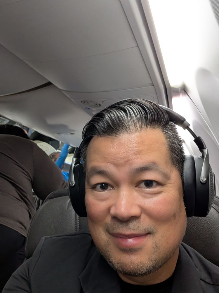 Pan-Pacific man with impeccable superhero hair, wearing headphones and sitting on a plane. 