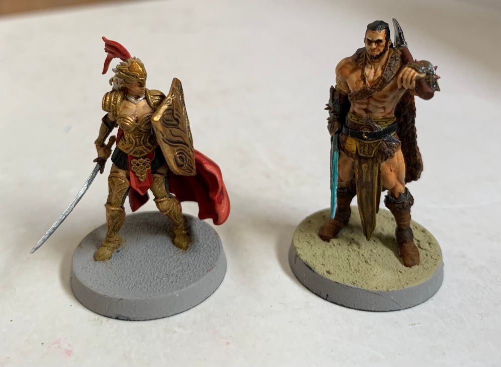 Painted gaming miniatures. On the left: an Amazon warrior in gold armour with a red cape, holding a longsword in her right hand. On the right: a barbarian in a brown cloak and loin cloth, holding a broadsword in his right hand, and with an axe resting on his left shoulder. 
