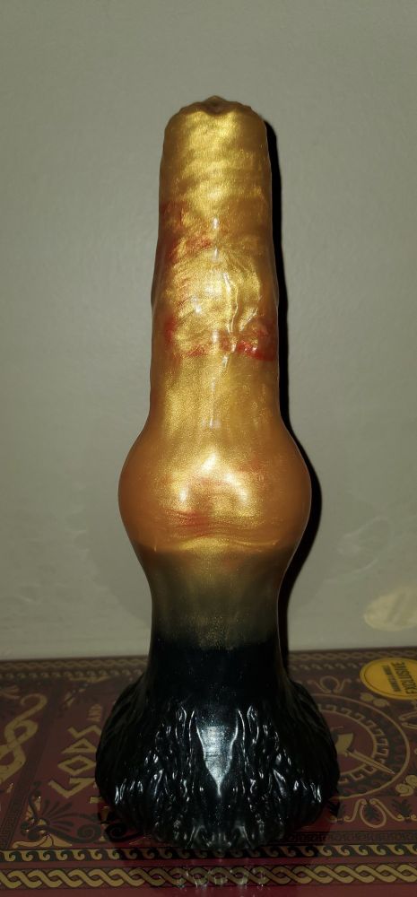 The image contains a dildo from https://www.fantasticocks.com. The dildo is Shadow the Dire Wolf, medium, "metallic gold" shaft with "shimmery black" base, added marbled "Leo red" to shaft. Front view.