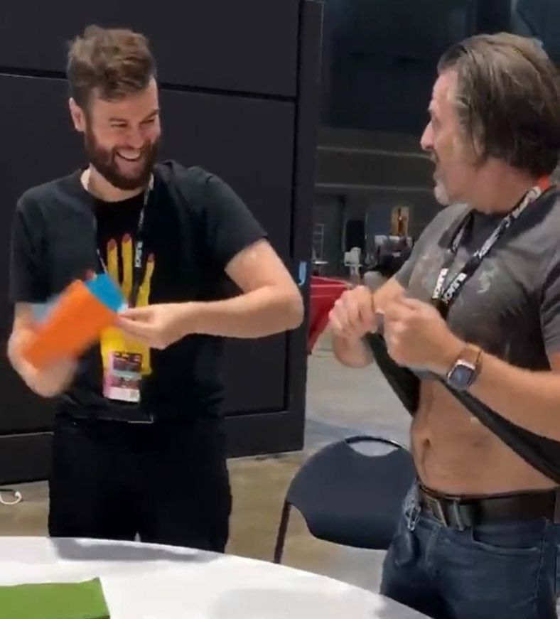 Con O'Neill and Nathan Foad backstage at a convention Con has his shirt raised while Nathan looks at him and smiles 