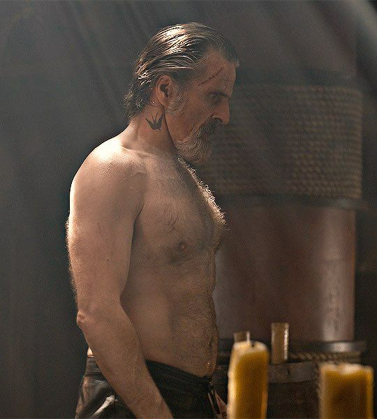 Con O'Neill as Izzy Hands he is shirtless standing sideways and surrounded by candles 