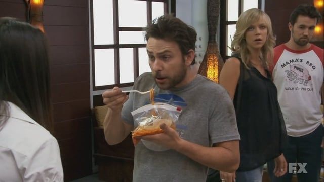 Charlie from It's Always Sunny in Philadelphia eating spaghetti from a zipper bag with a plastic fork. There's a big stain on the left arm of his T-shirt