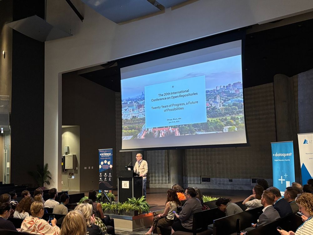 A man stands at a lectern and gives a presentation to a room of people. the text on the slide behind him reads "The 20th international conference on open repositories. twenty years of progress, a future of possibilities"  