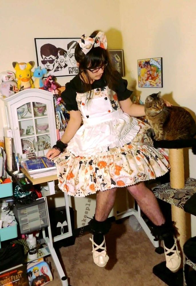 A light skin lolita sitting on a desk. To his left is a clear cabinet holding tea cups with pokemon plushies on top. To his right is a fluffy warm brown tabby cat. The lolita has dark brown hair with a large bow in the middle of his head. The fabric of the bow is the same as the jsk, an ivory base with an all over pattern of red yarn, black cats, and playing cards. He is wearing a black short sleeve blouse, black wristcuffs, and short black ruffle lace top socks. He is wearing ivory shoes with bows on them. Other background items include a Radkey poster, Hollow Knight artwork, and One Piece poster. There is a Naruto shippuden art pouch on the floor.