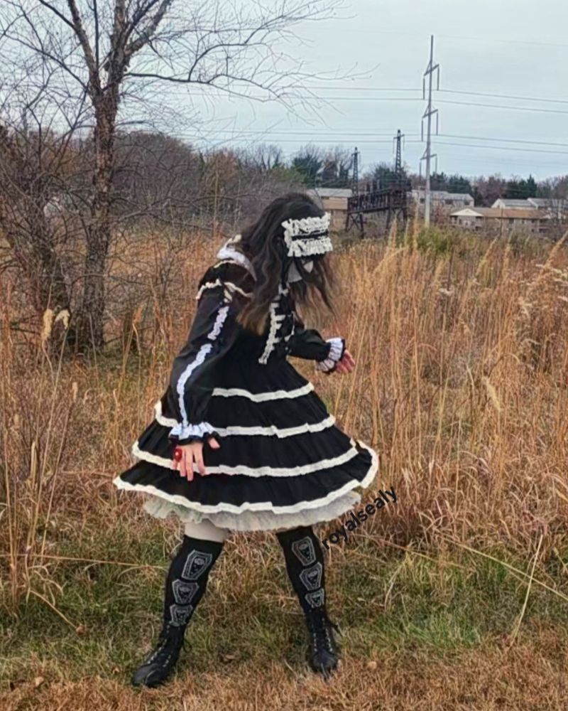 Royalsealy is center, standing in front of autumn brush. The sky is a gray blue, with buildings further back along the horizon. He is wearing the same black and white lolita coord but spinning, the bottom of his skirt full with the movement. He is looking down and away from the camera. His front most hand has a red rose ring on it.