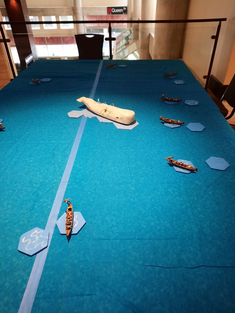View of a tabletop strategy game based on Moby Dick, depicting the ocean in blue felt, with tiny whaling ships dwarfed by a replica of Moby Dick at the center.