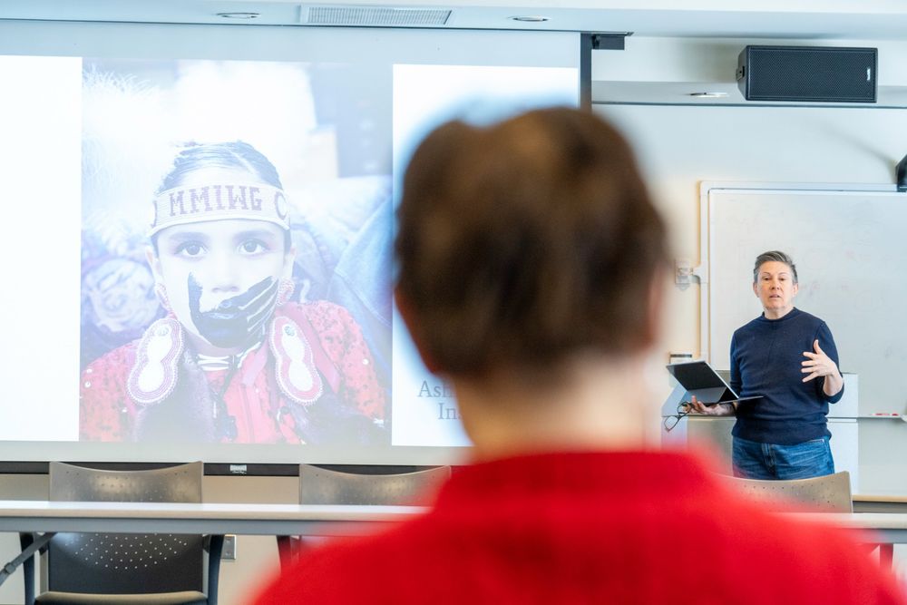 Professor El Chenier stands at front of room presenting. On screen is a photo of a young girl with a black hand painted on their mouth. She wears a headband that says "MMIWG." 