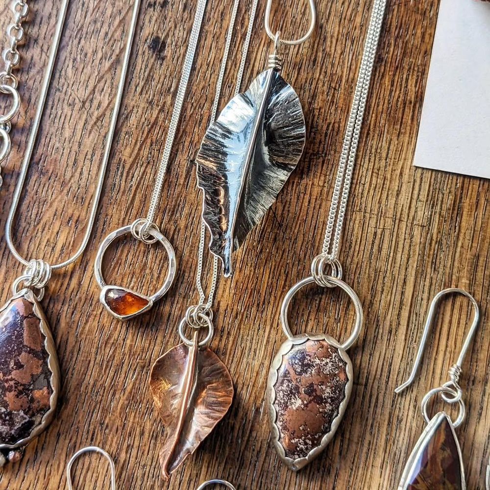 Close-up of other necklaces and earrings, with more silver and copper leaves, an amber stone mounted on a ring as part of a necklace, and other mottled brown stones in bezel settings with scalloped edges.