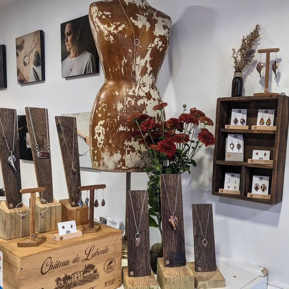 The corner of the shop counter in the LollaMac shop. There is a wooden wine box with necklaces and earring displayed on wooden stands. Other necklaces are displayed on wooden stands in front of a vase of red flowers and an old mannequin. Attached to the wall to the side of the counter is a wooden box with internal dividers, being used to display earrings on white cards.