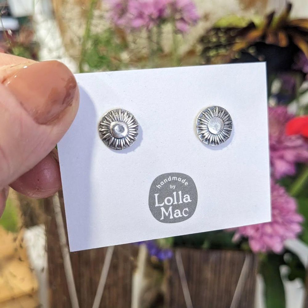 Two silver ear studs with impressions of sunflower blooms, mounted on a white card with the LollaMac logo. The card is being held by a thumb (the rest of the hand is out of frame). In the background are some unfocused flowers.