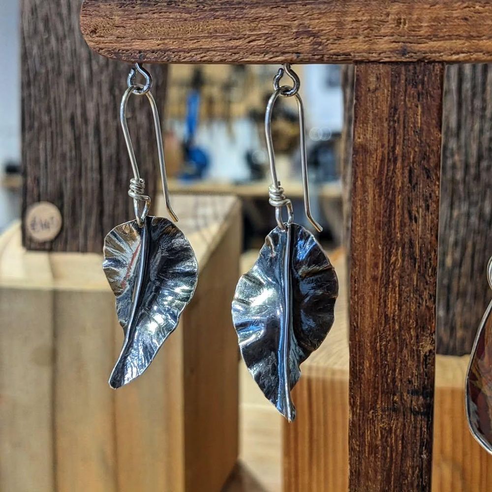Two silver earrings hanging from a wooden display stand. The earrings are fold-formed to look like leaves.