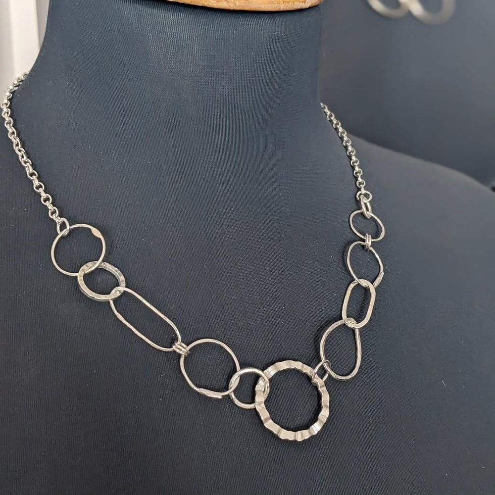 A silver necklace displayed on a black mannequin. The necklaces consists of connected silver hoops of various shapes and sizes, and features a distinctive hammered silver hoop in the middle.