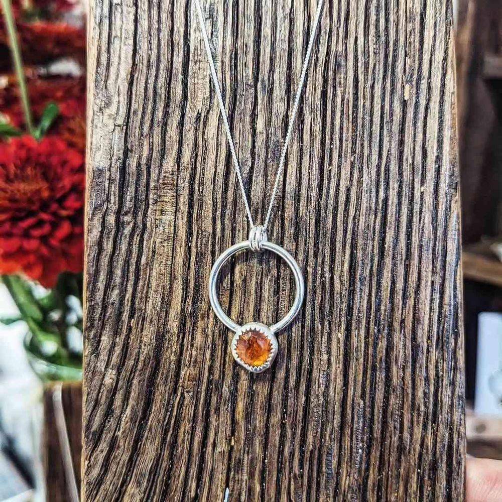 Necklace featuring a small hessonite garnet in a bezel setting attached to a silver ring. It is resting on a heavily grained wooden board.