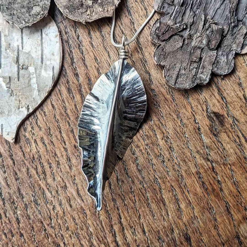 Silver necklace, formed to like a fallen leaf, resting on a heavily grained wooden surface