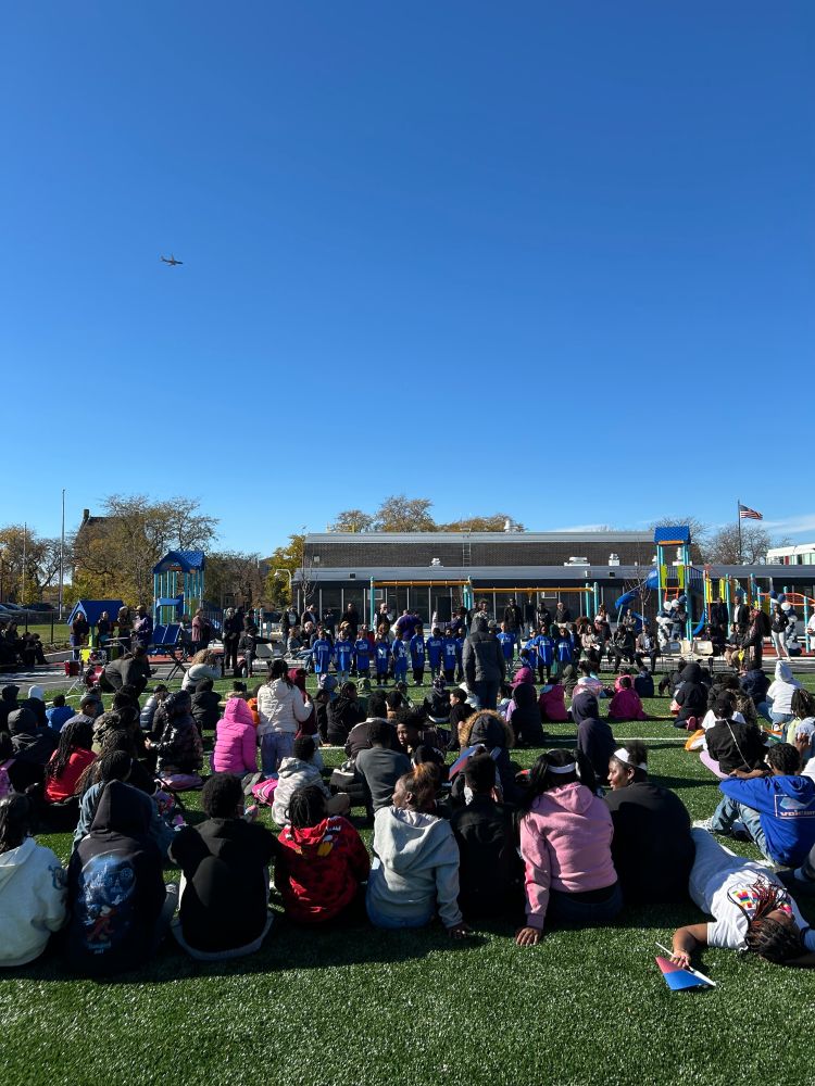 Crowd sitting on grass at a school during a sunny day, watching a performance by young students, with a clear blue sky, a playground, and the school building in the background.