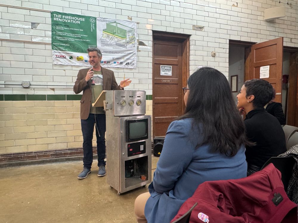 A person stands at a podium holding a microphone and addressing an audience. In the background is a tile wall displaying a banner with the heading "The Firehouse Renovation" and an architectural drawing of a building.