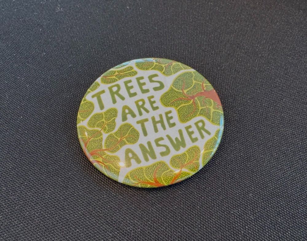 A button with the words "Trees Are the Answer" and decorative trees sits on a black tablecloth.