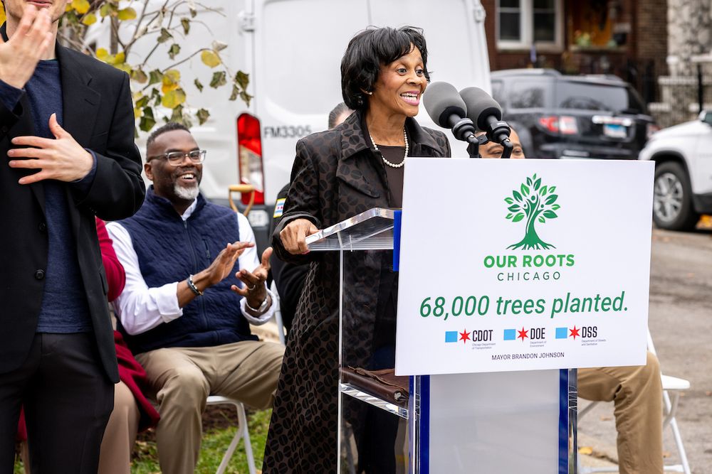 A person speaks at a podium bearing a sign with a logo of a tree and the words "Our Roots Chicago." Underneath it, the sign reads "68,000 trees planted" and includes the logos of CDOT, DOE, and DSS, as well as the name "Mayor Brandon Johnson." Behind the speaker, the Mayor smiles and claps.
