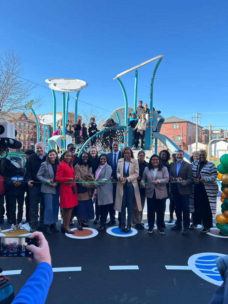 A group of individuals at a ribbon-cutting ceremony on a playground, with balloons, celebrating the opening of a new schoolyard.