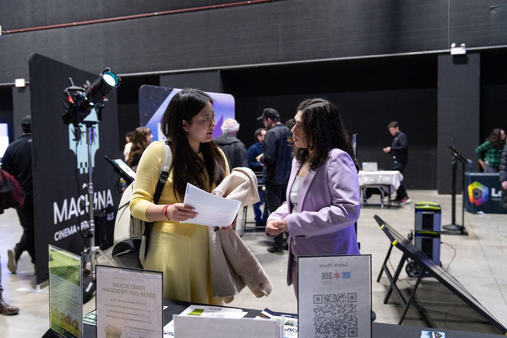 Two people engaged in conversation at an expo booth with signage and film industry equipment in the background.