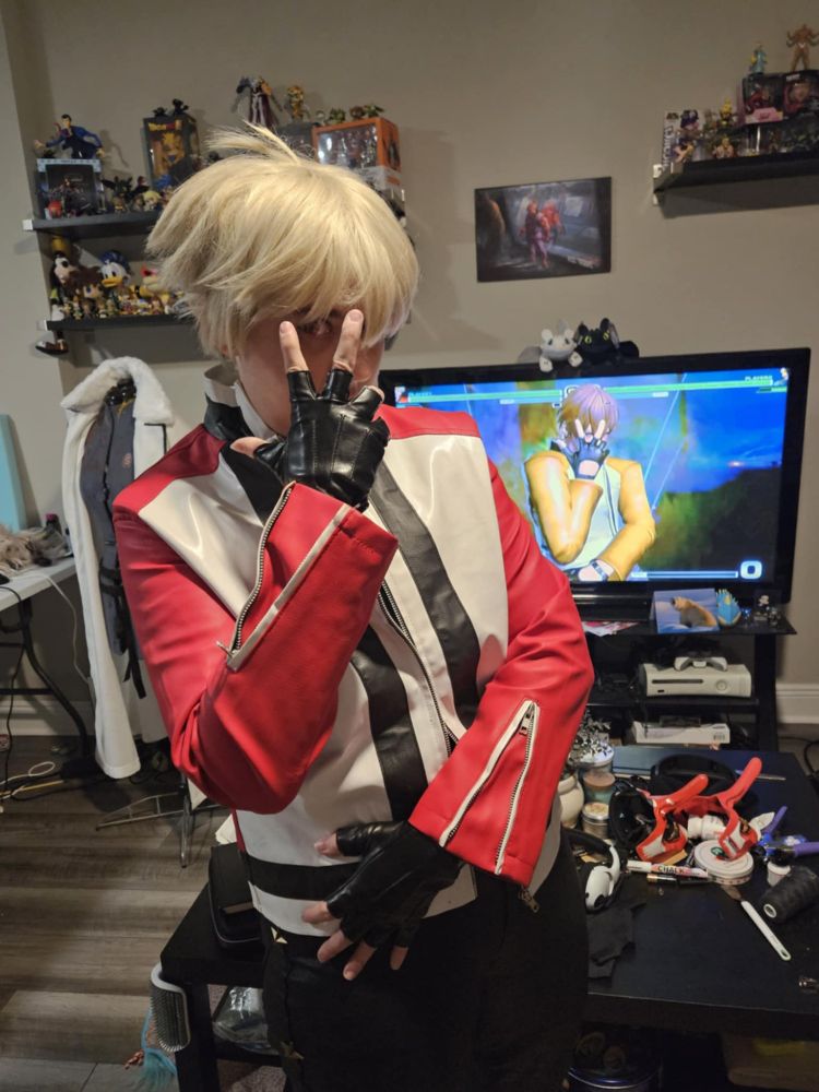 Me (SkywardKey, aka Sky) standing in my livong room dressed as Rock Howard from Fatal Fury/King of Fighters/SNK. I'm posing as his first pose for his Neo Deadly Wave Super Move - left arm crossing my body to rest on my right hip, and right arm raised with my hand covering most of my face. My right eye is visible between my index and middle fingers.
