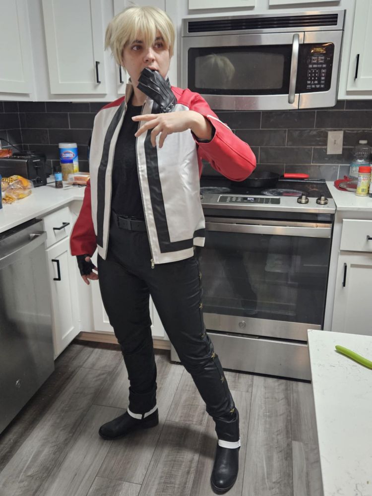 Me (SkywardKey, aka Sky) standing in my kitchen cosplaying Rock Howard from Fatal Fury/King of Fighters/SNK. I'm mimicking one of his victory poses - I'm looking off camera to the distance at a 3/4 view, legs apart, right arm relaxed at my side, left atm approximstely shoulder height mid-shake. I'm holding a black fingerless glove between my teeth.